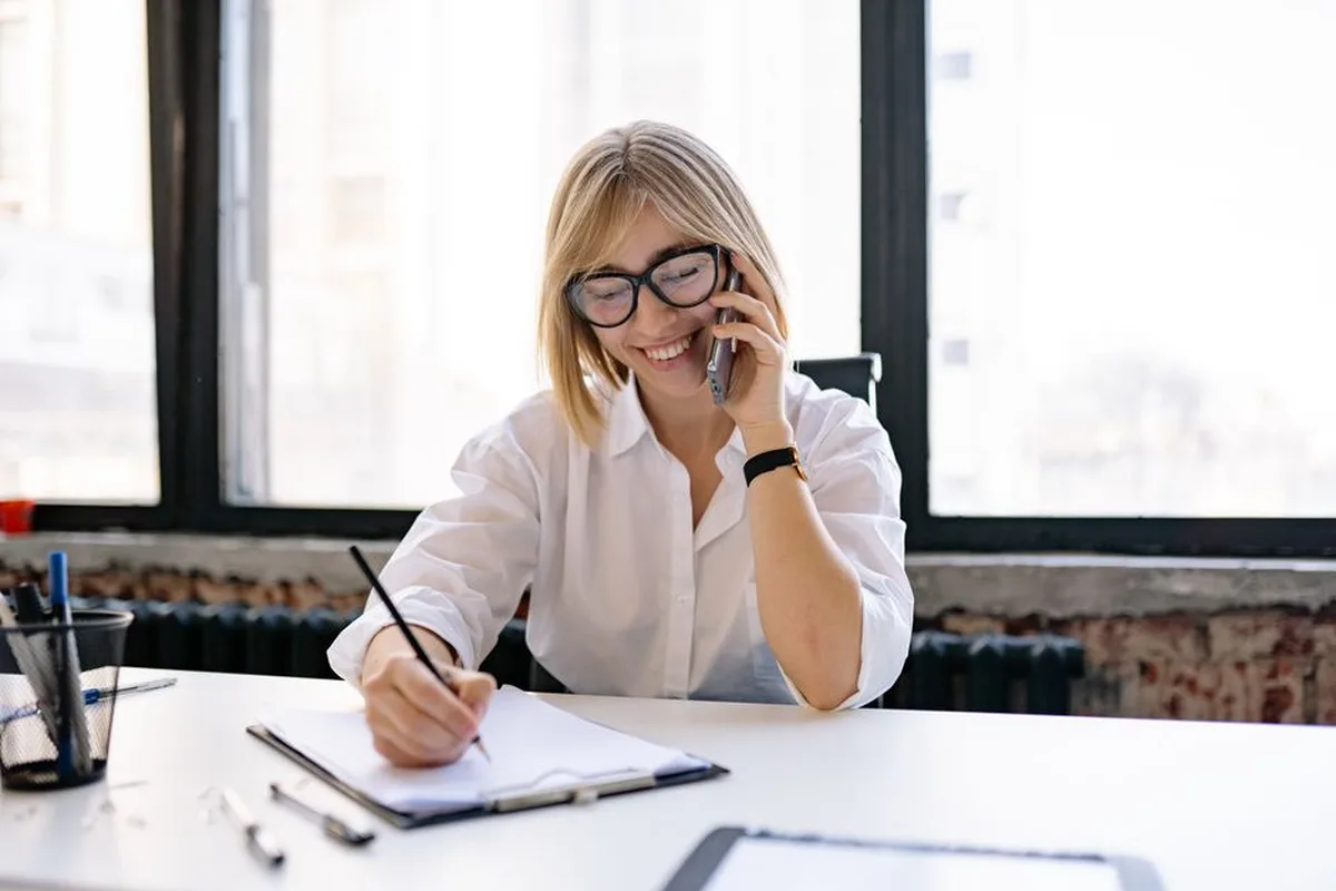 Une femme souriante prend des notes en écoutant un appel téléphonique à son bureau.