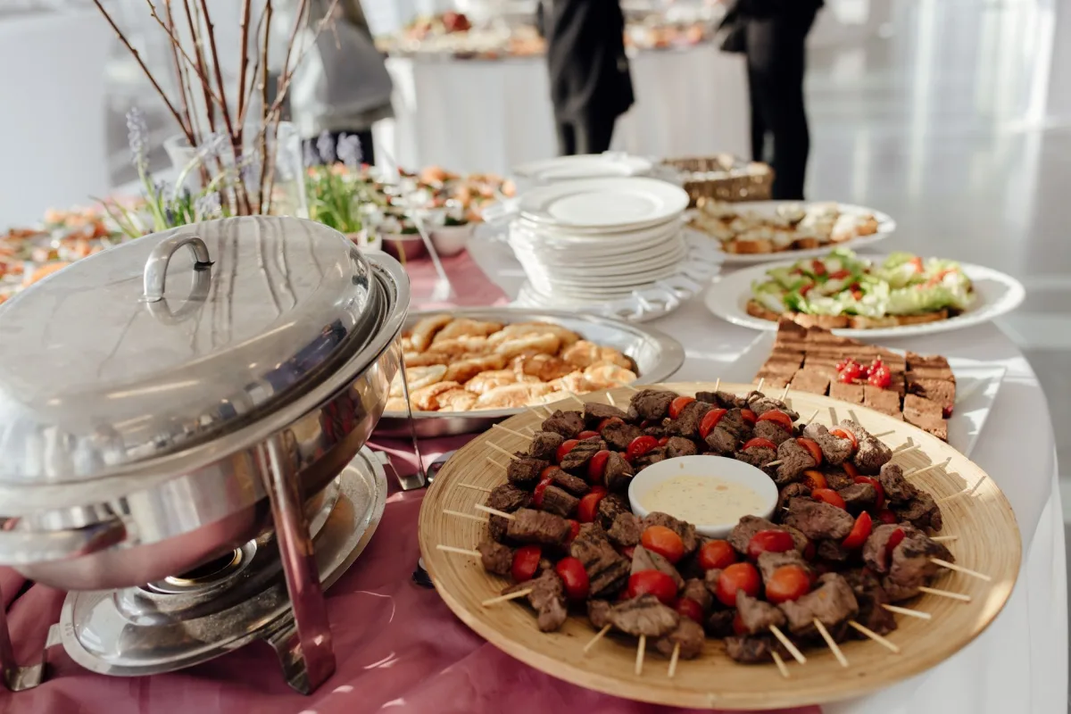 Buffet dressé sur une nappe rose : brochettes de viande et tomates cerises autour d'une sauce, salades, petits pains, desserts et piles d'assiettes à côté d'un chauffe-plat.