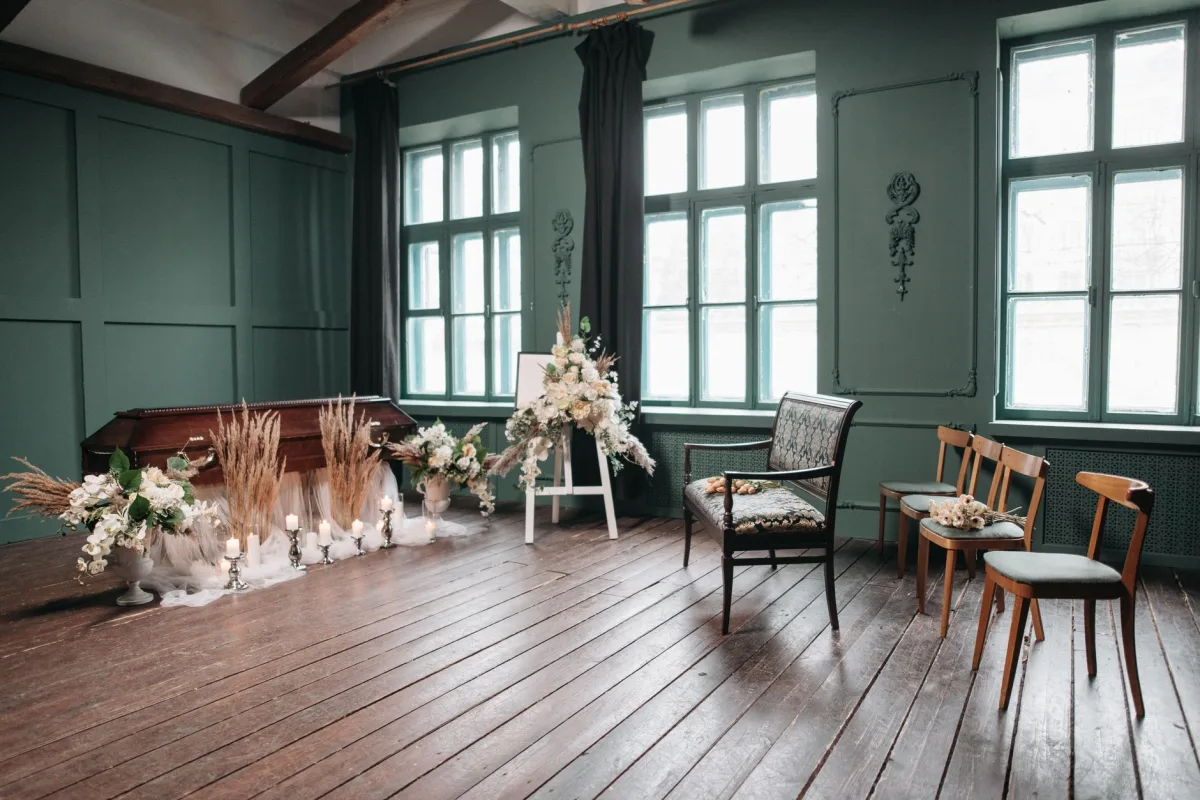 Salon funéraire sobre aux murs verts, avec un cercueil en bois, des compositions de fleurs blanches, des bougies et quelques chaises devant de grandes fenêtres.