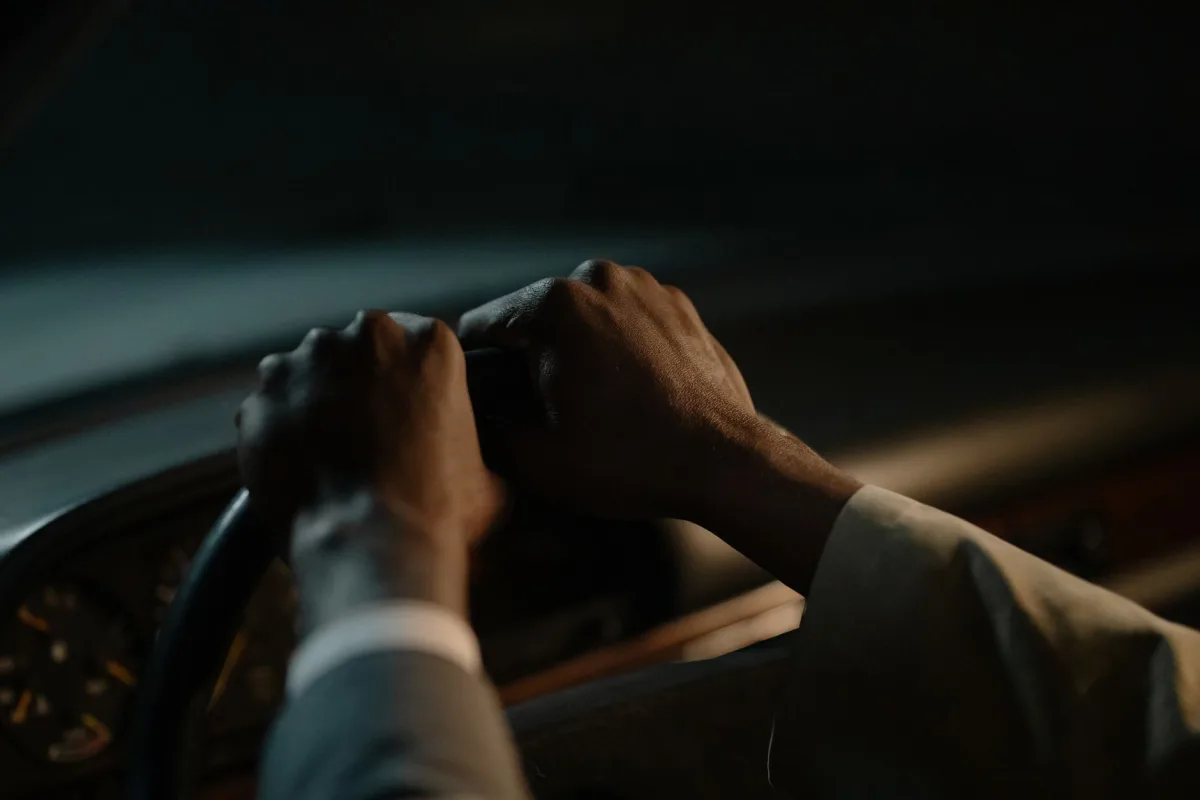 Les mains d'un chauffeur en veste de costume tiennent le volant dans l'habitacle sombre d'une voiture, le soir.